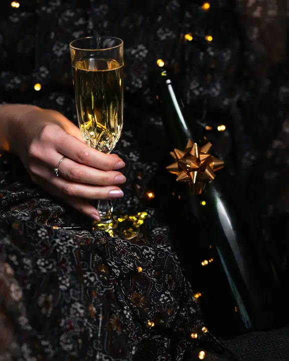 Tamna boca pjenušca sa zlatnom mašnicom, čaša šampanjca u ruci