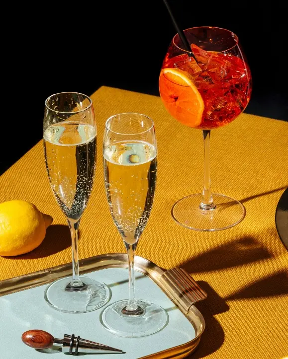 dvije čaše champagne na pladnju i čaša aperol spritza sa narančom i ledom pokraj