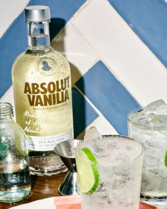 boca Absolut Vanilia Flavored Vodka 0,7l u elegantnoj boci okruženoj čašama s koktelima, ledom i kriškama limete na ljetnoj barskoj sceni